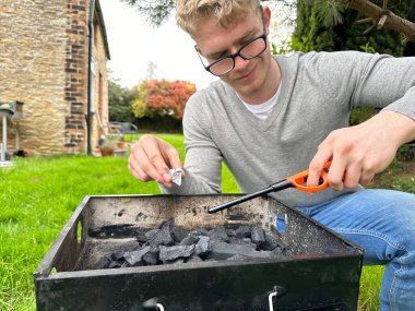 Yakışıklı genç adam kömür ızgarasında ateş yakıyor, bahçesinde bbq için hazırlanıyor.