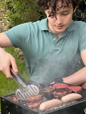 Genç bir adamın portatif ızgarada sosis ve hamburger pişirirken çekilmiş dikey görüntüsü.