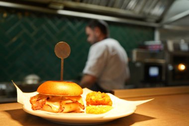 Lokantadaki sıcak lambanın altındaki hamburgerlere odaklan. Arka planda açık planlı mutfakta aşçı olarak çalış.