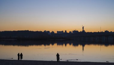 Kyiv, Ukrayna - 20 Mart 2025: Dnipro nehrinin kumsalında dinlenen insanlar, Ukrayna 'nın mücadelesinin sembolü olan Ukrayna' nın Ukrayna 'daki tam ölçekli Rus işgalinin (24 Şubat 2022) ve Nat' ın bir parçası olan Ukrayna Anıtının manzaralı manzarasına karşı