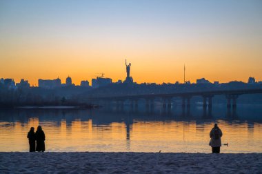 Kyiv, Ukrayna - 20 Mart 2025: Dnipro nehrinin kumsalında dinlenen insanlar, Ukrayna 'nın mücadelesinin sembolü olan Ukrayna' nın Ukrayna 'daki tam ölçekli Rus işgalinin (24 Şubat 2022) ve Nat' ın bir parçası olan Ukrayna Anıtının manzaralı manzarasına karşı