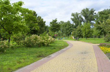 Bereketli yeşil ağaçlar ve çalılar, huzurlu bir park ortamında renkli bir oyun alanına giden dolambaçlı asfalt bir yol çerçevesi oluşturarak, çocukların dinlenmesi için mükemmel bir yer sunuyor.