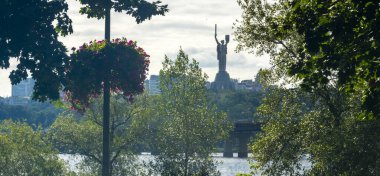 Tomurcuklanmış ağaçlar ve canlı çiçek sepetleri ikonik Anavatan Anıtı 'nı çerçeveliyor Kyiv siluetine karşı yükseliyor dingin Dinyeper nehri alttan akıyor.