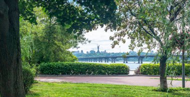 Dnieper Nehri ve Kyiv şehrinin silueti ile çevrili kaldırımlı bir yol üzerinde ağaçlar ve çalılarla çevrili yeşil bir park. Arka planda ana Ukrayna anıtı bulunuyor.