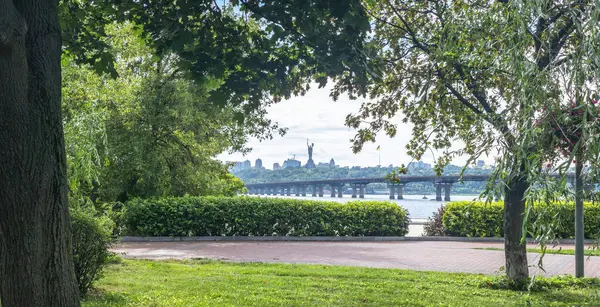 Dnieper Nehri ve Kyiv şehrinin silueti ile çevrili kaldırımlı bir yol üzerinde ağaçlar ve çalılarla çevrili yeşil bir park. Arka planda ana Ukrayna anıtı bulunuyor.