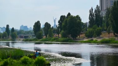 Dnipro nehrinin kıyısında yemyeşil bir bitki örtüsü var. Orada bir balıkçı, ana Ukrayna anıtı ve Kyiv kentinin zeminine doğru yol alıyor.