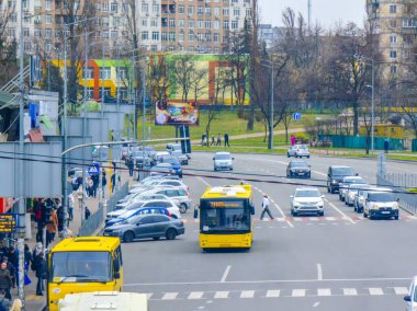 Kyiv, Ukrayna - 20 Mart 2025: Sarı toplu taşıma otobüsü park edilmiş araçlar, yayalar otobüs durağında bekliyor ve bulutlu bir yay altında konut binaları