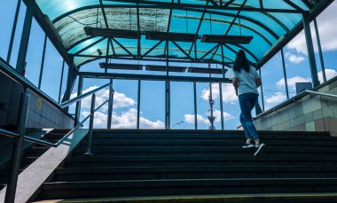 Kyiv, Ukrayna - 25 Temmuz 2025: Kyiv metro istasyonunda, şeffaf mavi bir çatı altında, parlak mavi gökyüzü ve istasyonun pencerelerinden görünen beyaz bulutlarla merdivenleri tırmanan genç bir kadın