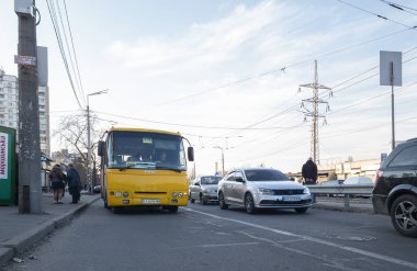 Kyiv, Ukrayna -6 Mart 2025: Ukrayna 'da gündüz vakti bir sokakta giden sarı otobüs, otobüs durağında bekleyen arabalar ve yayalar, Doğu Avrupa' da kentsel ulaşımı gösteriyor