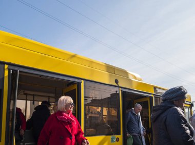 Kyiv, Ukrayna - 6 Mart 2025: Aralarında kırmızı ceketli zarif bir kadının da bulunduğu yolcular, açık mavi gökyüzü olan güneşli bir günde Ukrayna 'dan sarı bir toplu taşıma otobüsüne biniyorlar.