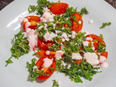 Dilimlenmiş domates ve taze roka, üzerinde kremalı yoğurt sosu bulunan açık mavi bir tabakta karıştırılarak basit ama canlı ve sağlıklı bir yemek veya yan yemek oluşturur.