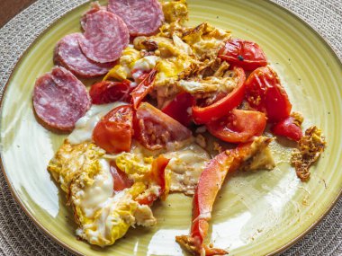 Çırpılmış yumurta, domates, salam ve kırmızı dolma biber, ketojenik, protein zengini bir yemeği betimleyen bir masa paspasına yerleştirilmiş sarı bir tabakta servis edilir.