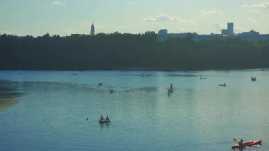 Ukrayna 'nın başkenti Kyiv' de, güneşli bir yaz gününde arka planda şehrin ufuk çizgisi ile birlikte birçok insan kano yapıyor, kano yapıyor ve kürek çekiyor.