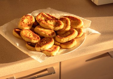 Syrniki olarak bilinen altın esmer peynir krepleri mutfak tezgahındaki kağıt havlunun üzerinde beyaz bir tabağa dizilmiş, güneş ışığının sıcacık parıltısıyla yıkanmış.