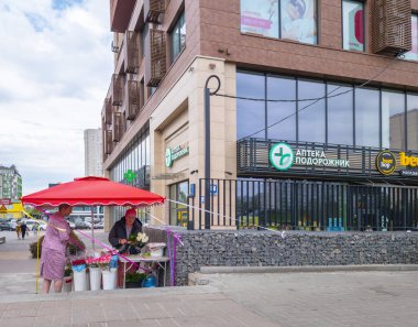 Kyiv, Ukrayna - 20 Temmuz 2025: İki kadın Ukrayna 'daki bir sokak pazarında kırmızı şemsiye altında çiçek buketleri düzenliyor, modern binalar ve canlı toplum yaşamlarıyla çevrili.