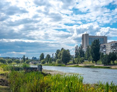Bereketli yeşil bitki örtüsü Dnipro Nehri bulutlu bir gökyüzünün altında huzur içinde akıyor. Uzaktan yükselen ikonik Ukrayna Ana Anıtı pitoresk bir Kyiv sahnesi oluşturuyor.