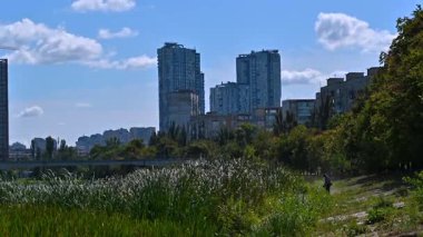 Şehir bakım işçisi nehir kenarındaki çimenleri köprü ve şehir binalarıyla biçiyor, mavi bulutlu gökyüzü altında canlı yeşillik ve kentsel altyapı ile yaz günü manzarasını yakalıyor.