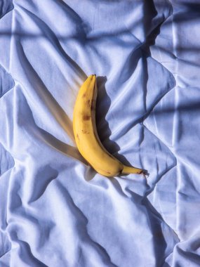 Yumuşak mavi yatak çarşafları üzerinde dinlenen olgun bir muz yakındaki bir pencereden süzülen güneş ışığıyla aydınlatılmış, huzurlu ve davetkar bir sahne yaratıyor.