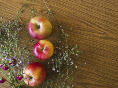 Üç olgun kırmızı elma, narin mor ve pembe çiçeklerle çevrili ahşap bir masada dinlenerek doğal güzelliği kutlayan büyüleyici ve kırsal bir hayat yaratıyor.