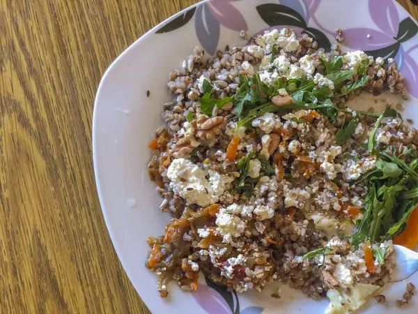 Karabuğday lapası, haşlanmış sebze, roka, feta peyniri, ceviz ve ayçekirdeği ile ahşap bir masada servis edilen tabakta sağlıklı ve lezzetli bir yemek seçeneği sunuyor.