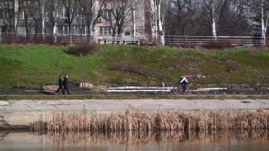 Unidentified cyclist riding a bicycle along a concrete embankment next to a river on a spring day, with other people walking and resting on benches in the background green park