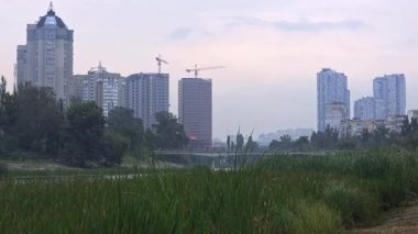 Modern cityscape with residential high rise buildings and construction cranes standing behind a lush green park with a river. Urban development concept shows the contrast between nature and city life