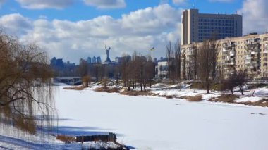 Kyiv, Ukraine - 10th Januaryl, 2025: Winter cityscape of Kyiv, Ukraine, featuring a frozen river covered in snow, residential buildings, and the iconic Motherland Monument visible against a cloudy blue sky on a sunny day