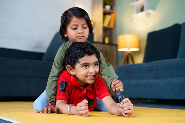 Young siblings kids fighting for remote control while watching tv or television at home - concept of childhood lifestyle, siblings quarrel and technology