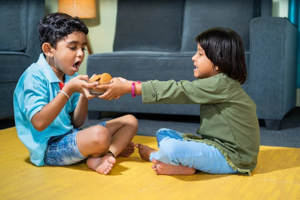 Young siblings kids fighting for snacks or biscuit while sitting on floor at home - concept of childhood lifestyle, relationships and weekend holidays.