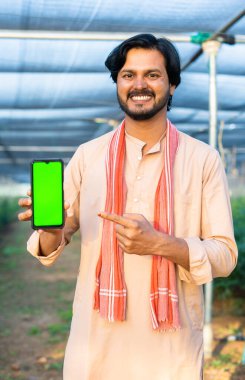 vertical shot of Happy smiling young farmer showing green screen mobile phone by pointing finger by looking camera at greenhouse - concept of agriculture promotion, advertisement and app promotion.