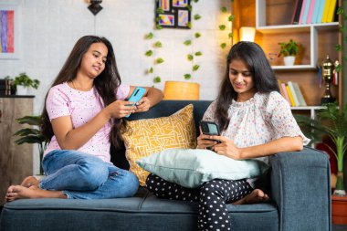 siblings busy using mobile phone while sitting on sofa at home - concept of social media addiction, cyberspace and relationship