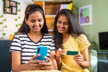 Happy smiling young siblings making online payment for shopping on mobile phone by using credit card at home - concept of e commerce, online banking and bonding.
