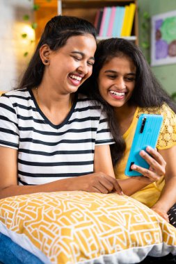 vertical shot of laughing sibling girls while using mobile phone on sofa at home - concept of entertainment, social media and internet connection