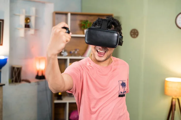 sweet excited young man with VR or virtual reality goggles doing workout by using joystick at home - concept of metaverse, health care and technology