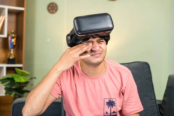 Young man rubbing eyes due to over use of VR or virtual reality goggles or headset at home while on sofa - concept of overuse of technology, addiction and healthcare