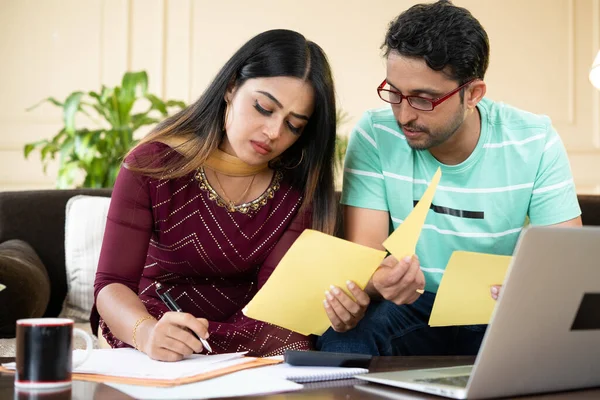 Serious young indian couple calculating expenses or monthly bills in front of laptop while sitting at home - concept of investment, budget and financial planning.