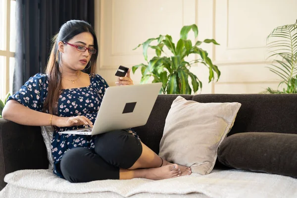 Young woman making online payment on laptop using credit card while sitting on sofa at home - concept of e-commerce shopping, bill payment and shopaholic.