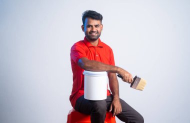 Confident painter sitting by holding paint brush with bucket by looking camera - concept of home improvement, job and renovation service