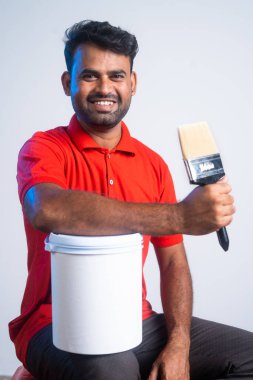 Vertical shot of smiling painter holding paint brush and bucket by looking at camera while sitting at workplace - concept of home renovation, successful and employment.