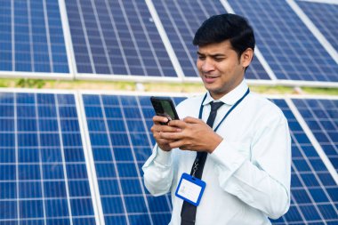 Young engineer busy using mobile phone in font of solar panel at power plant - concept of maintenance service, technology and renewable energy