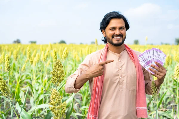 Portrait shot of Happy Indian farmer showing money by pointing finger while looking camera at farmland with copy space - concept of earning, agriculture profit and successful investment.
