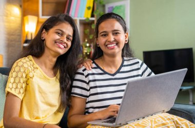 Happy smiling siblings girl using laptop by looking at camera at home - concept of technology, learning and bonding.