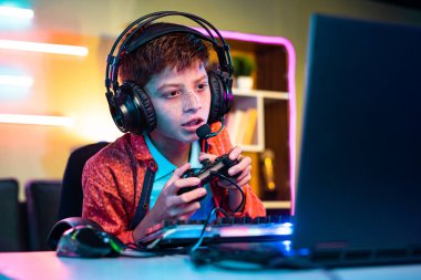 Teenager kid playing online video gameon laptop by talking on headphones at home - concept of pro gamer, tournament and live streaming