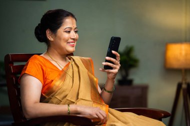 Middle woman in sare buy using mobile phone on chair at home - concept of technology, social media sharing, internet