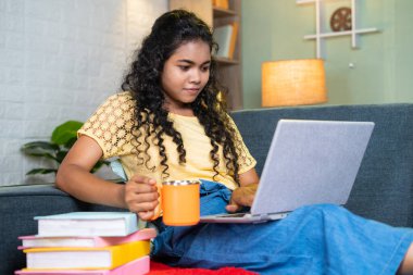 İnceleme sırasında dizüstü bilgisayar kullanırken kahve ya da çay içen Hintli genç bir kız evde kanepede kitaplarla otururken - ferahlatıcı, serbest çalışan ve online alışveriş kavramı.