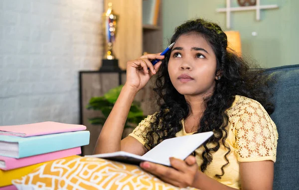 thoughful girl while reading book at home while sitting on sofa during exam preparation - concept of hard working, exam preparation and education.