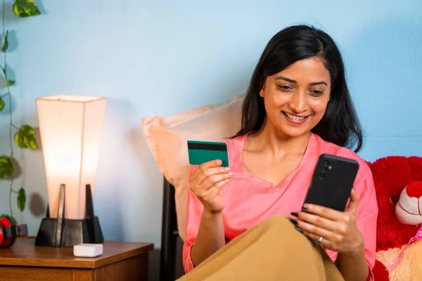 Happy young girl making online payment using credit card on mobile phone during sales or offers at while sleeping - concept of online secure transaction, midnight festive discounts