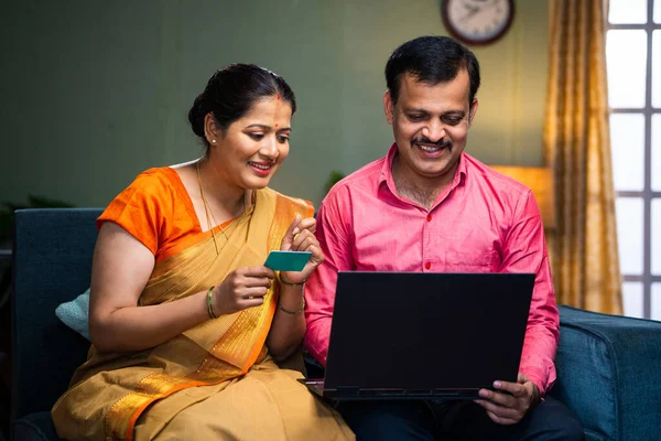 Happy smiling indian couple making online payment for shopping using credit card on laptop on sofa at home - concept of e commerce, financial or banking and family.