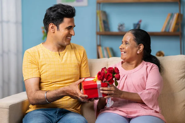 快乐的印度成年儿子在家里坐在沙发上送红玫瑰送给年长的母亲 母亲节的概念 感情和感情 — 图库照片