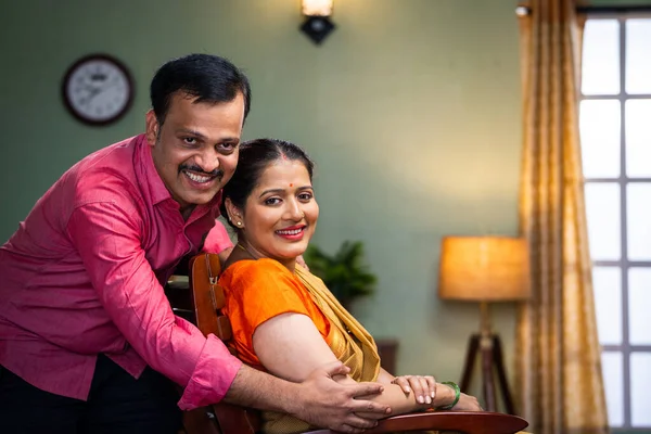 portrait of Happy smiling middle aged couple looking at camera while sitting on chair at home - concept of successful relationship, family bonding and supportive husband